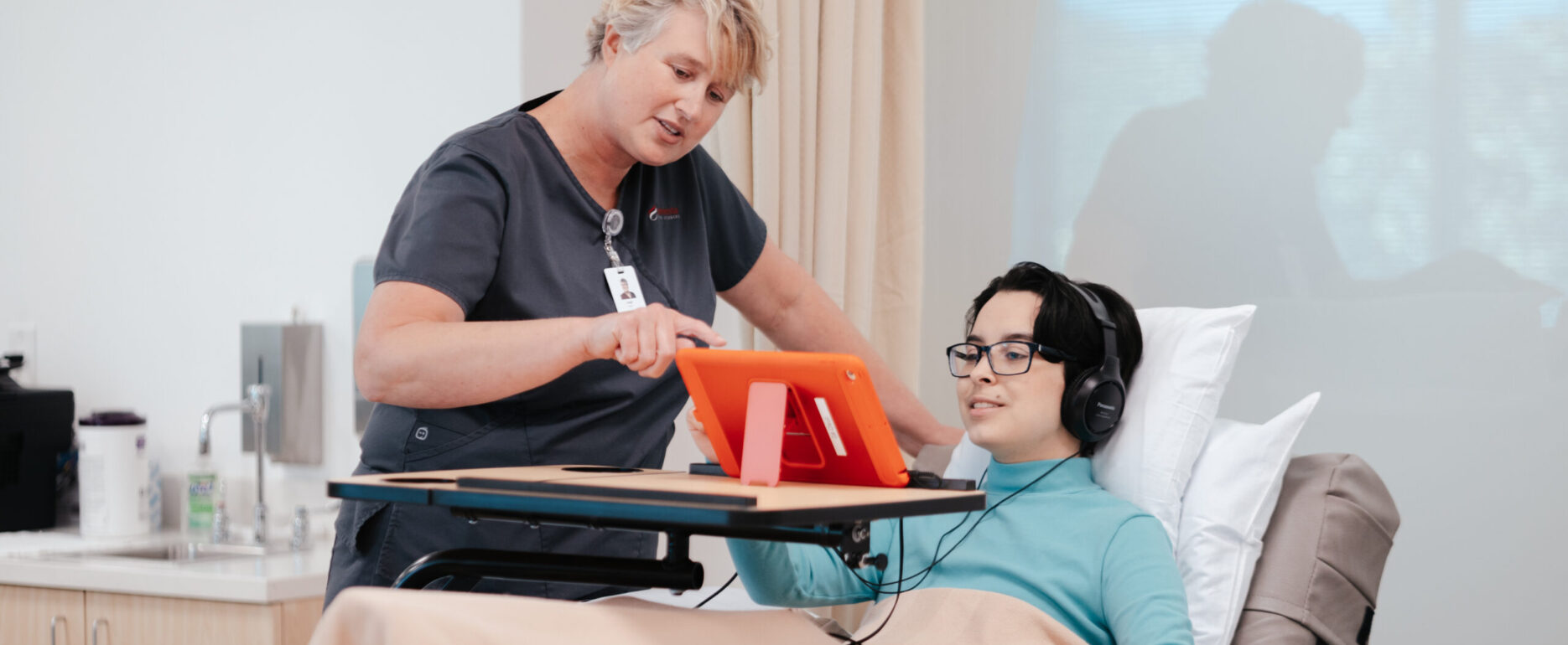 A nurse with short blonde hair assists a blood donor, who is reclining in the donation chair, with operating the provided iPad donors can use during their blood donation.