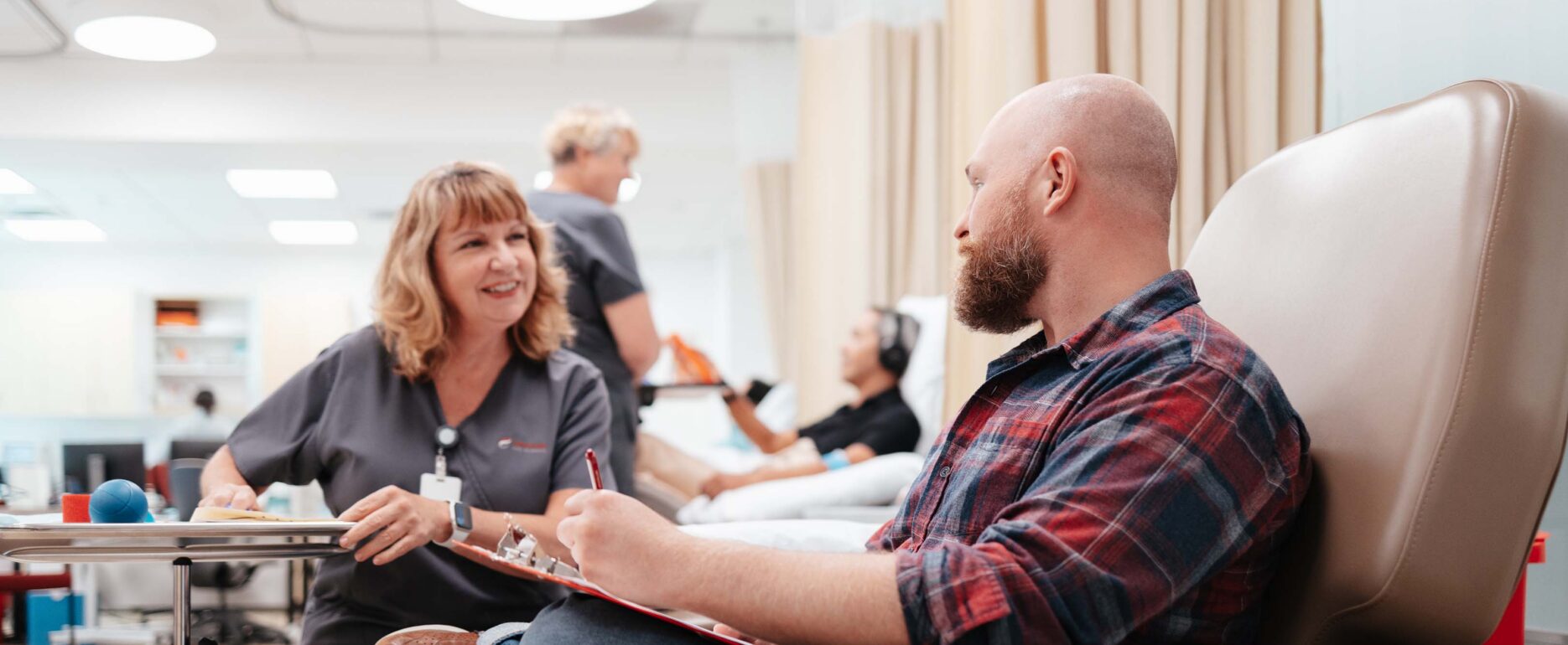 A male donor with a shaved head and short beard sits in the Canventa office across from a female nurse with shoulder-length blonde hair.